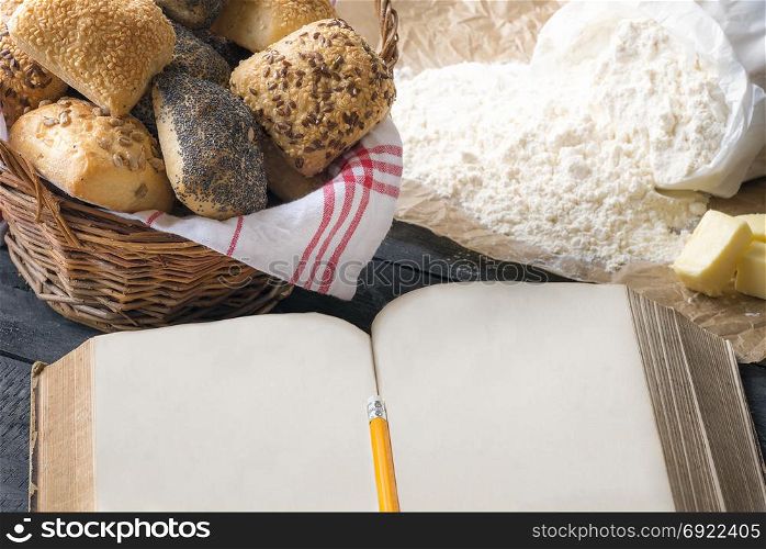 Antique open book with space for text, surrounded by fresh homemade bread rolls and baking ingredients. A concept for baking, cooking, recipe.