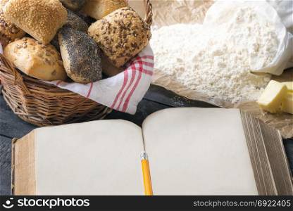 Antique open book with space for text, surrounded by fresh homemade bread rolls and baking ingredients. A concept for baking, cooking, recipe.