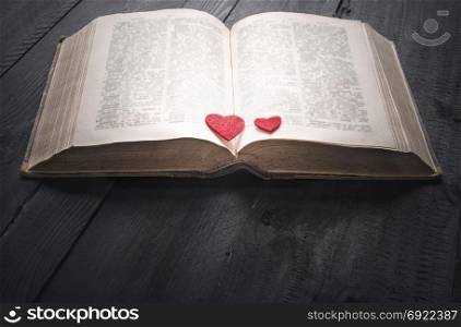 Antique open book with two red hearts on its pages, sitting on a black wooden background. A concept for the love of reading, learning, education or as a love story.