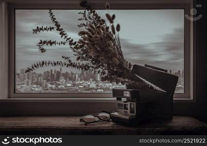 Antique polaroid camera, sunglasses and Dried flowers in old wooden box by the glass wall with city view. Vintage tone style, Copy space for text, Selective focus.