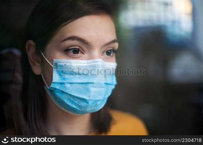Anxious depressed and stressed young woman wearing medical face mask,in home isolation looking outside window, uncertain future due to global Coronavirus COVID-19 pandemic crisis, stay at home concept