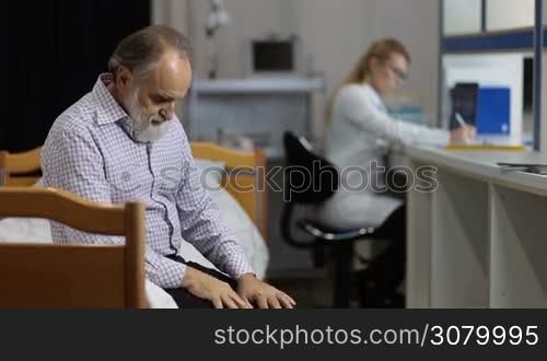 Anxious senior male patient sitting on examination table waiting for medical results in clinic with blurry female doctor making notes on background. Worried retired man expecting to hear medical results from physician while sitting on hospital ward.