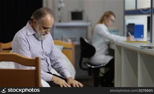 Anxious senior male patient sitting on examination table waiting for medical results in clinic with blurry female doctor making notes on background. Worried retired man expecting to hear medical results from physician while sitting on hospital ward.