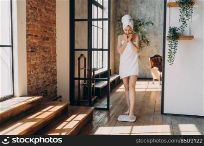 Anxious slim caucasian woman taking step onto scale to measure her weight. Girl wrapped in towel after bathing has time at spa. Young woman checking weight after shower. Fitness and wellness concept.. Anxious slim caucasian woman wrapped in towel taking step onto scale to measure her weight.