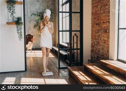 Anxious slim caucasian woman taking step onto scale to measure her weight. Girl wrapped in towel after bathing has time at spa. Young woman checking weight after shower. Fitness and wellness concept.. Anxious slim caucasian woman wrapped in towel taking step onto scale to measure her weight.