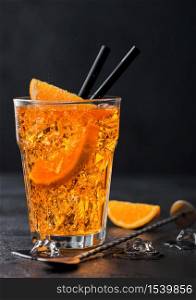 Aperol spritz summer cocktail in highball glass with orange slices and bar spoon on black background with ice cubes.