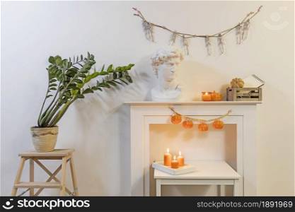 Apollo&rsquo;s plaster head. Zamioculcas plant in clay pot on stool. A garland of skeletons hangs on wall. A garland of plastic pumpkins hangs from fireplace. The interior is decorated for Halloween.