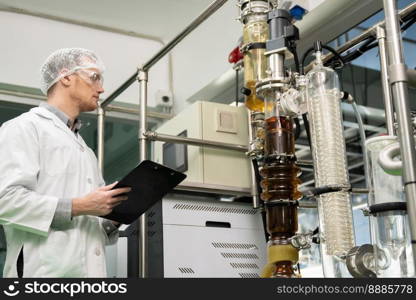 Apothecary scientist using a clipboard and pen to record information from a CBD oil extractor and a scientific machine used to create medicinal cannabis products.. Apothecary scientist recording data from CBD oil extractor in laboratory.