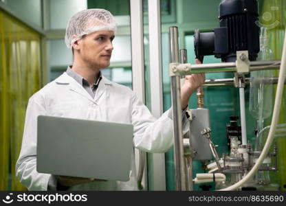 Apothecary scientist using laptop to record information from a CBD oil extractor and a scientific machine used to create medicinal cannabis products.. Apothecary scientist recording data from CBD oil extractor in laboratory.