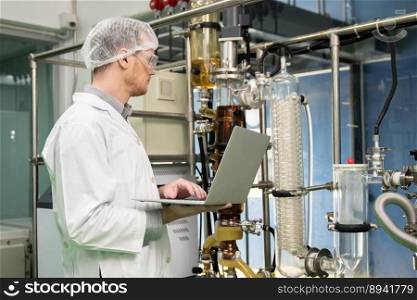 Apothecary scientist using laptop to record information from a CBD oil extractor and a scientific machine used to create medicinal cannabis products.. Apothecary scientist recording data from CBD oil extractor in laboratory.