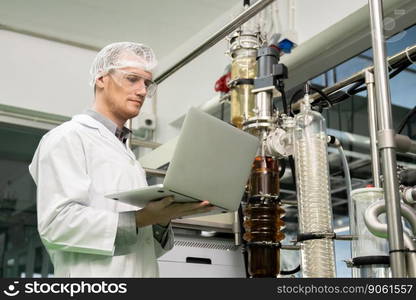 Apothecary scientist using laptop to record information from a CBD oil extractor and a scientific machine used to create medicinal cannabis products.. Apothecary scientist recording data from CBD oil extractor in laboratory.