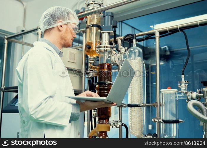 Apothecary scientist working with laptop near CBD oil extractor and a scientific machine used to create medicinal cannabis products.. Apothecary scientist working with laptop near CBD oil extractor in laboratory.