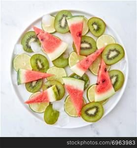 Appetizing juicy pieces of kiwi, lime and watermelon with ice cubes in a plate on a gray marble background. Flat lay. Plate with pieces of fresh kiwi fruit, watermelon and lime on a gray marble background. Flat lay