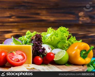 Appetizing still life. Fresh cherry tomatoes, lettuce leaves, mushrooms, green basil, cheese on wooden board. Vegetables isolated on brown wall.