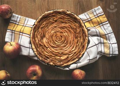 Apple pie decorated in shape of a rose flower, in a tray, on kitchen towel, surrounded by apples fruits, on vintage table. Above view of tatsy apple pie in rustic settings.