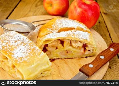 Apple strudel with icing sugar, apples, tea strainer, knife on a wooden boards background