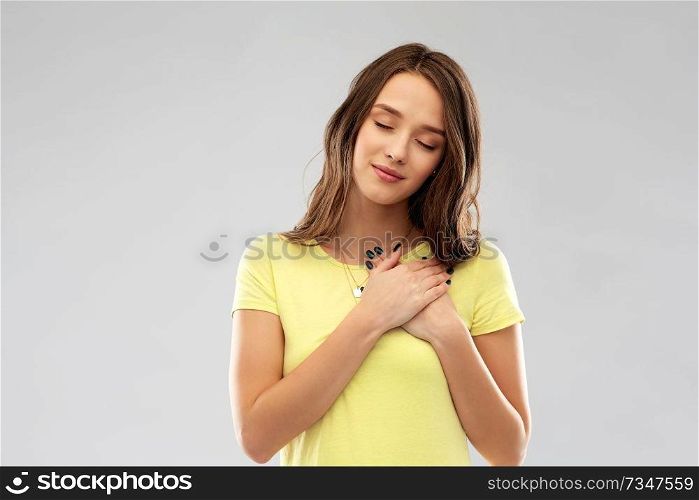 appreciation, valentine’s day and sincere emotions concept - thankful young woman or teenage girl with closed eyes in yellow t-shirt holding hands on chest or heart over grey background. smiling teenage girl holding hands on heart