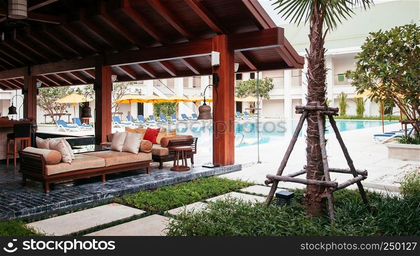APR 26, 2013 Phuket, Thailand - Resort style couch, wood table with fabric cusion and pillow under wooden pavilion in green garden next to swimming pool. Warm asian atmosphere