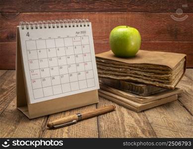 April 2022 - spiral desktop calendar with old book or journal and apple against rustic barn wood background, time and business concept
