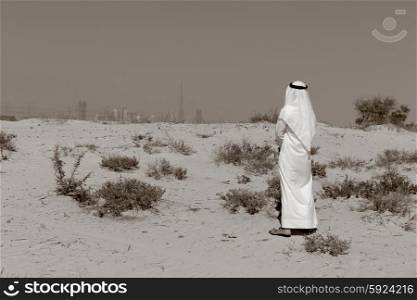 Arab man in national dress stands in the desert and looks at the city of Dubai. Toned.. Arab man in national dress stands in the desert and looks at the city of Dubai