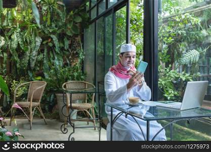 Arab muslim businessmen wear white traditional clothing in hand use smart phone and open laptop on coffee glass table