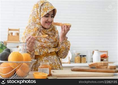 Arab Muslim cute kid girl wear hijab looking and smelling after spreading jam on sliced whole wheat bread for breakfast with fresh milk, oranges and many fruits for juices on kitchen table at home