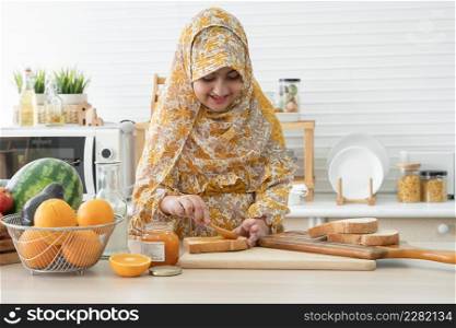 Arab Muslim cute kid girl wear hijab smiling holding spoon and spreading jam on sliced whole wheat bread for breakfast with fresh milk, oranges and many fruits for juices on kitchen table at home