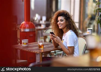 Arab woman in an urban bar at her smartphone. . Young arabic woman smiling and sitting in an urban bar in the street looking at her smartphone. Arab girl in casual clothes drinking a soda outdoors.