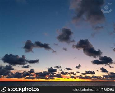 Arch of small dark dramatic clouds in the clear sunset sky colored in blue, pink, yellow and orange, over the Baltic sea, with small silhouettes of boat and lighthouses on the horizon