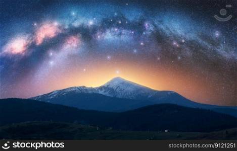 Arched Milky Way over the beautiful mountains with snow covered peak at night in summer. Colorful landscape with bright starry sky with Milky Way arch, snowy rocks, hills, orange light. Space. Nature 