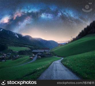 Arched Milky Way over the rural mountain road in summer in Italy. Beautiful night landscape with starry sky, milky way arch, winding road in mountain village, hills, green meadows and buildings. Space