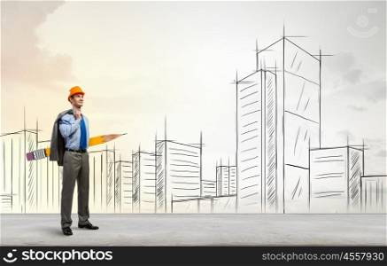 Architect designer with huge pencil. Young male engineer in hard hat with big pencil and buildings sketches at background