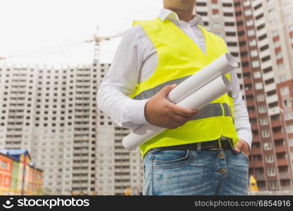 Architect in safety vest posing with blueprints at new buildings