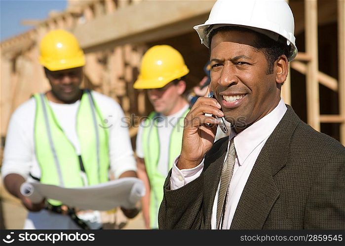 Architect using mobile phone and two construction works looking at blueprints
