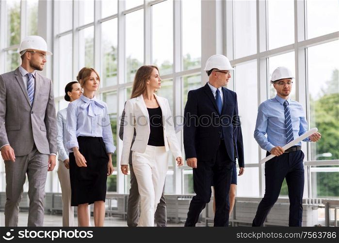 architecture and construction business concept - happy group of architects in helmets with blueprint walking along office building. business team in helmets walking along office