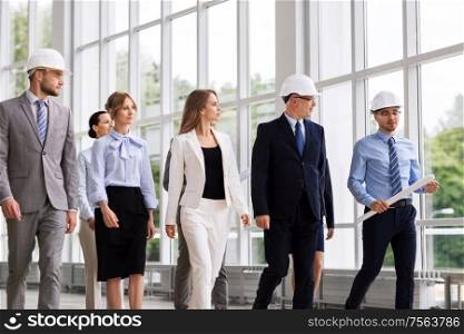 architecture and construction business concept - happy group of architects in helmets with blueprint walking along office building. business team in helmets walking along office