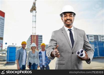 architecture, building business and people concept - smiling indian male architect in helmet with blueprints giving hand for handshake over construction site background. architect making handshake at construction site