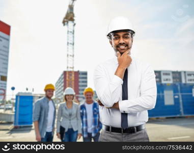 architecture, building business and people concept - smiling indian male architect in helmet over construction site background. indian architect in helmet over construction site