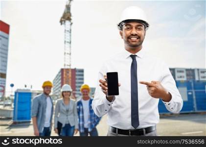 architecture, building business and people concept - smiling indian male architect in helmet showing smartphone over construction site background. architect showing smartphone at construction site