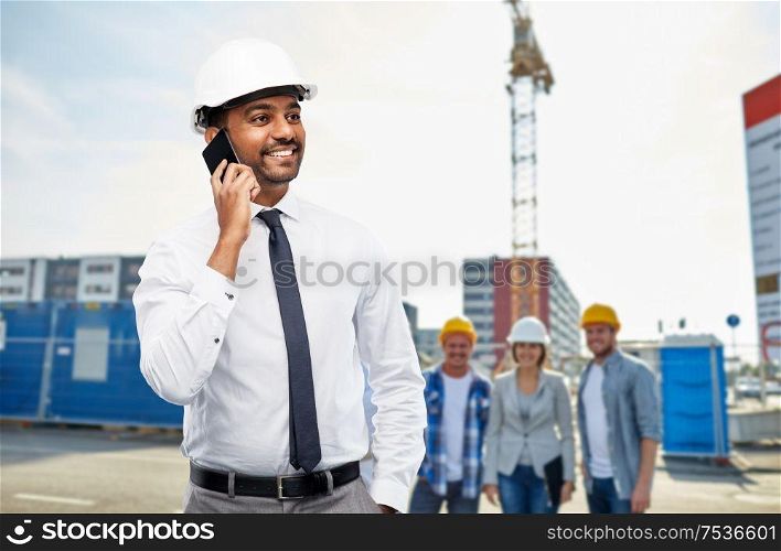 architecture, building business and people concept - smiling indian male architect in helmet calling on smartphone over construction site background. architect calls on smartphone at construction site