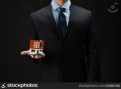 architecture, building, construction, real estate and property concept - close up of businessman or real estate agent holding house or home model on palm over black background