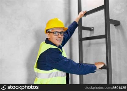 architecture, construction and building concept - happy smiling male builder or worker in helmet and safety west climbing ladder or stairs. builder or worker in helmet climbing ladder