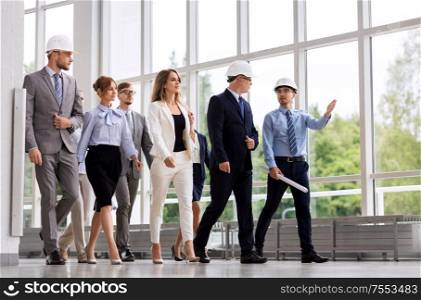 architecture, construction business and building concept - happy group of architects in helmets with blueprint walking along office building. business team in helmets walking along office