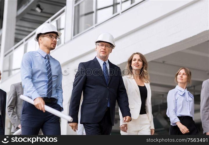 architecture, construction business and building concept - happy group of architects in helmets with blueprint walking along office building. business team in helmets walking along office