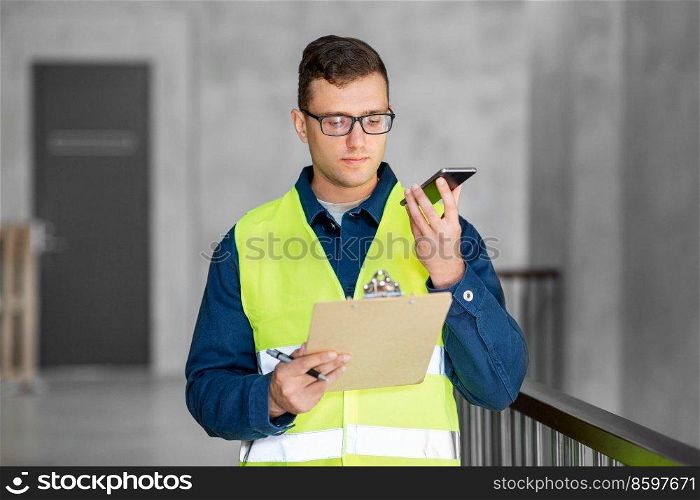 architecture, construction business and building concept - male architect in safety west with clipboard using voice command recorder on smartphone at office. male architect with clipboard calling on phone