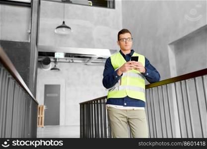 architecture, construction business and building concept - male supervisor in safety west with smartphone at office. male supervisor with smartphone at office