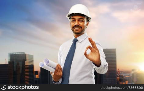 architecture, construction business and building - smiling indian architect or businessman in helmet with blueprints showing ok hand sign over sunset in tokyo city background. architect with blueprints showing ok in tokyo