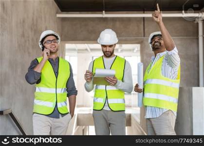 architecture, construction business and people concept - group of male architects in helmets with smartphone, blueprint and clipboard working at office building. male architects in helmets working at office