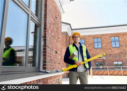 architecture, construction business and people concept - happy smiling male architect or builder in helmet and safety west with level and blueprint on site. male architect in helmet with level on site