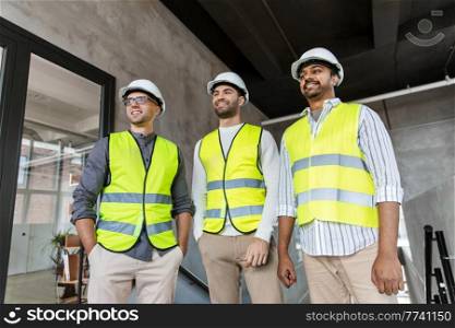 architecture, construction business and people concept - happy smiling male architects in helmets and reflective safety wests at office. architects in helmets and safety wests at office